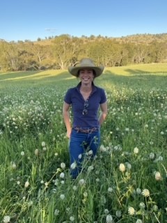 woman dressed in jeans and blue shirt wearing a hat standing in a green field with flowers